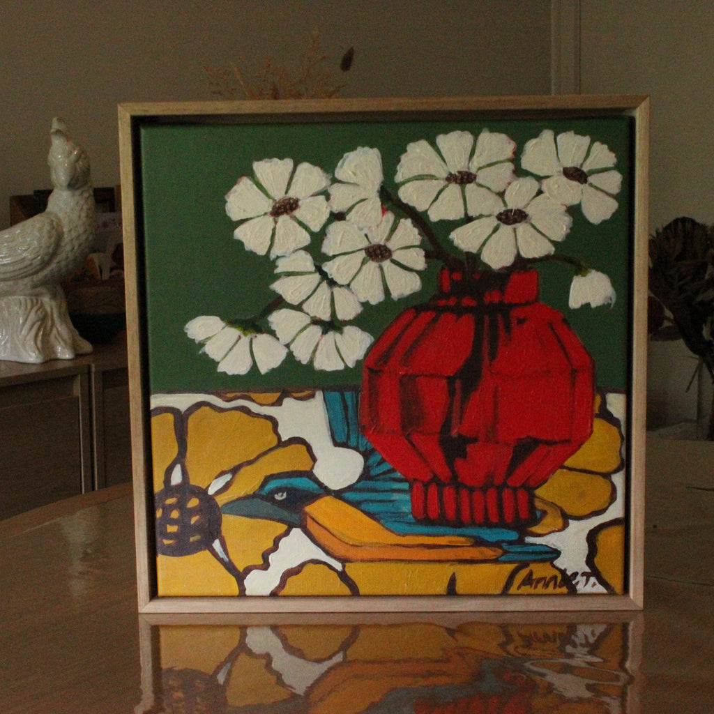 A framed square art print featuring a stylized still life with white flowers, a red vase, and a blue bird on a yellow background, displayed on a reflective table in an indoor setting.