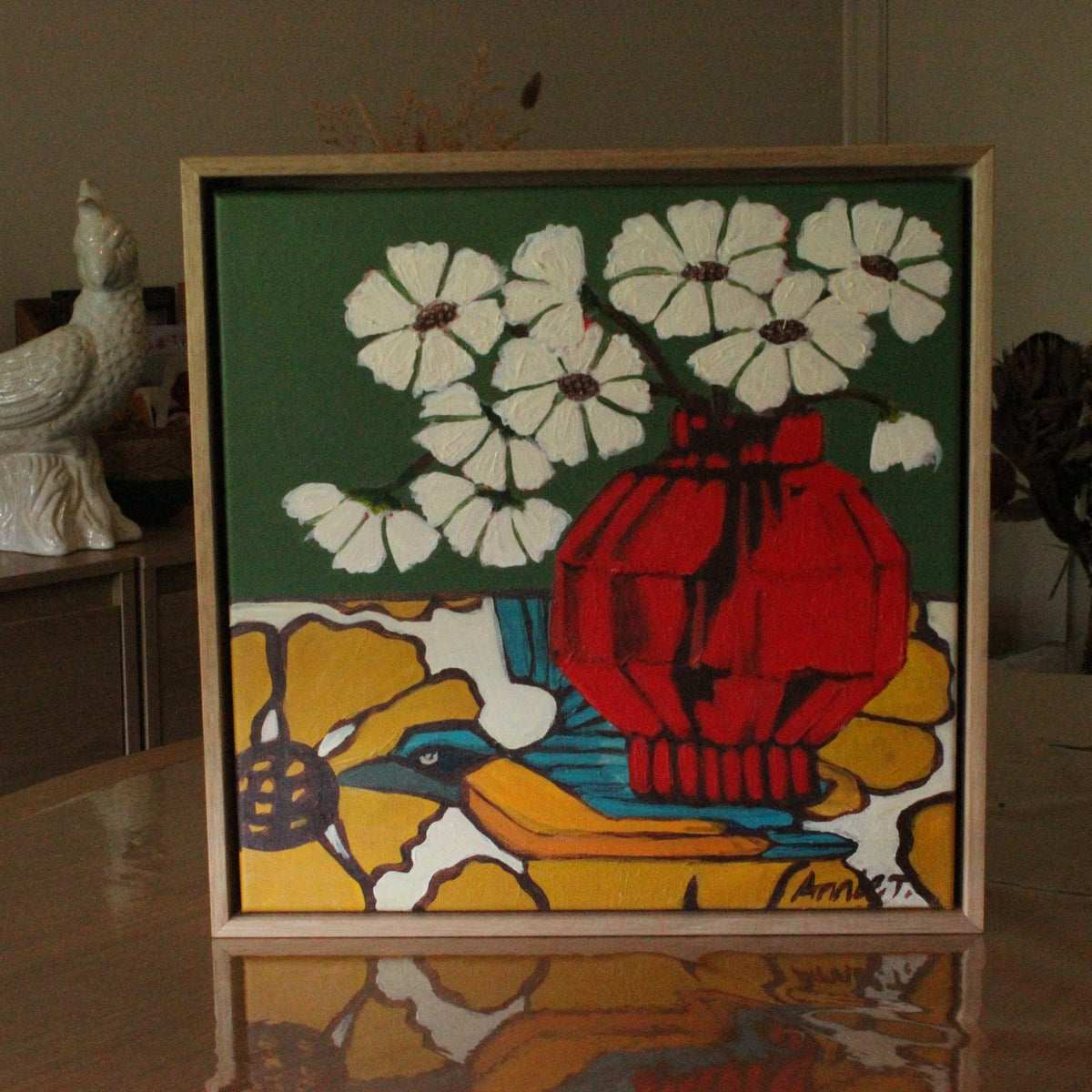 A framed square art print featuring a stylized still life with white flowers, a red vase, and a blue bird on a yellow background, displayed on a reflective table in an indoor setting.