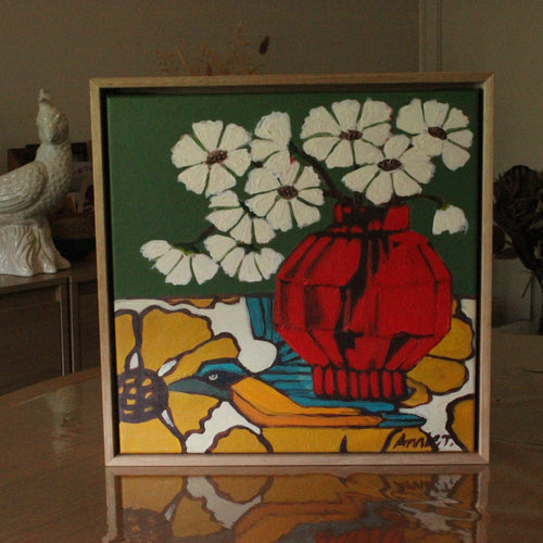 A framed square art print featuring a stylized still life with white flowers, a red vase, and a blue bird on a yellow background, displayed on a reflective table in an indoor setting.